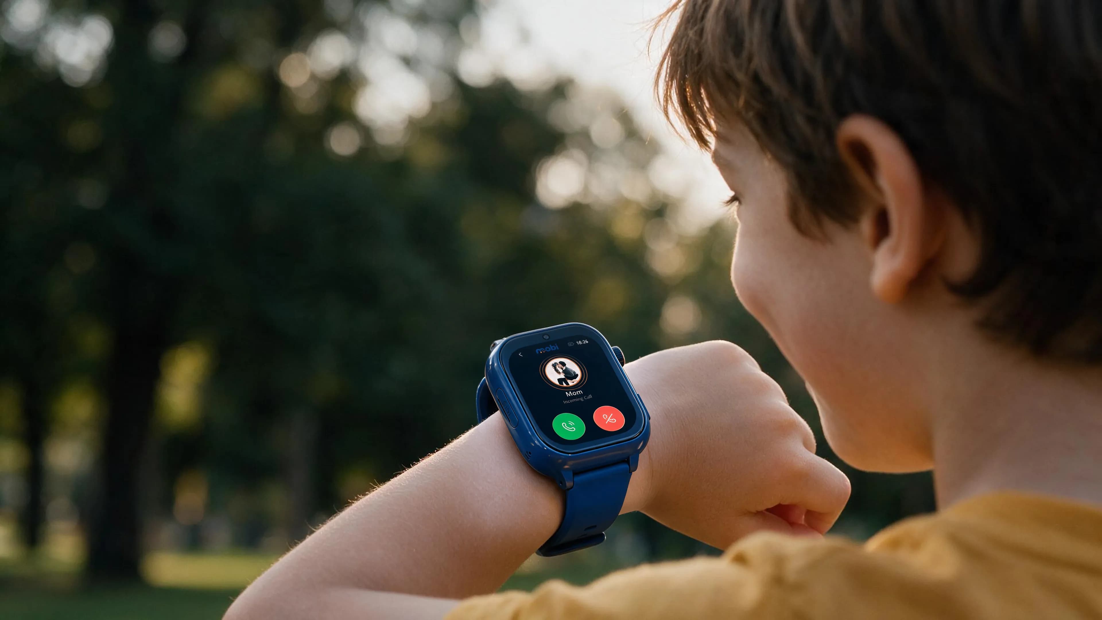 Seen over a child's shoulder in a park: a navy Mobi watch on their wrist showing an incoming call from Mom.