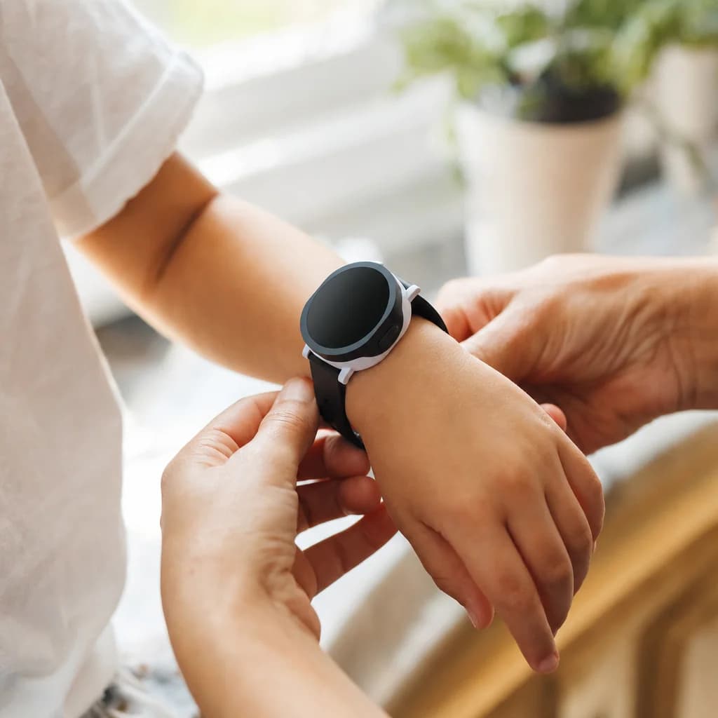 A parent helping their child fasten a Mobi smartwatch on their wrist. Warm afternoon light, plants in the background.